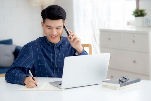 Young asian man talking phone and work from home with laptop computer, freelance male writing on notebook and speak on smartphone, stay home, domestic life, business and communication concept.