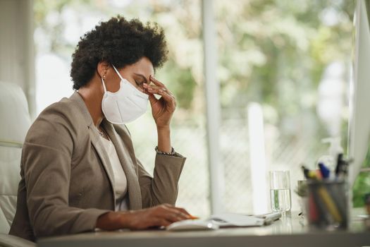 Shot of a stressed African businesswoman with protective N95 mask sitting alone in her office and working on computer during COVID-19 pandemic.