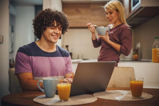 Shot of a happy couple spending their morning time together and talking in the kitchen. They are using laptop and eating breakfast before work.