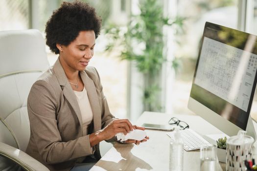 Shot of an attractive African businesswoman sitting alone in her office and using antiseptic gel to disinfect computer mouse during corona virus pandemic.