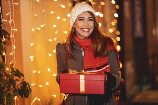 Cheerful young woman with red present box is having fun in the city street at Christmas time.
