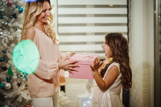 Shot of a cute little girl and her attractive mom are sitting on the floor, by the white Christmas tree and opening their Christmas presents.