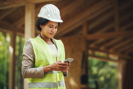 Shot of an African female architect using a smartphone and checking construction site of a new wooden house. She is wearing protective workwear and white helmet.