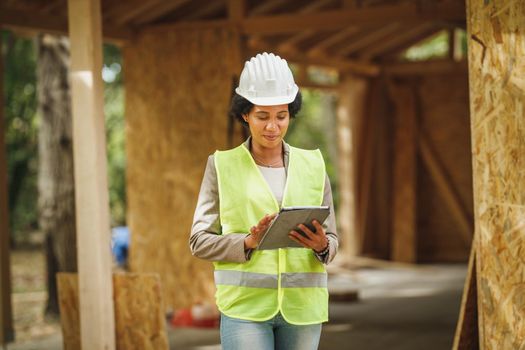 Shot of an African female architect using a digital tablet and checking construction site of a new wooden house. She is wearing protective workwear and white helmet.