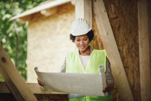 Shot of an African female architect checking plans at the construction site of a new wooden house. She is wearing protective workwear and white helmet.