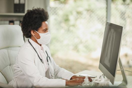 Shot of an African female doctor sitting alone in her consulting room and looking X-ray on computer during COVID-19 pandemic.