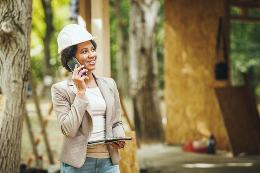 Shot of an African female architect with white helmet using a smartphone and checking construction site of a new wooden house.
