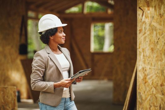 Shot of an African female architect with white helmet using a digital tablet and checking construction site of a new wooden house.