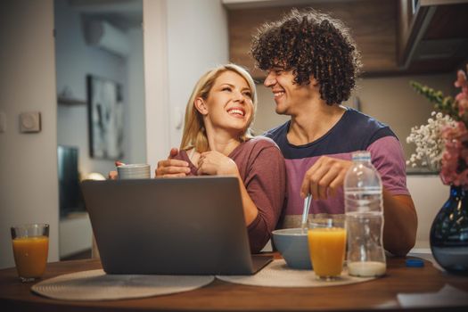 Shot of a happy couple using laptop together and eating breakfast before work in the kitchen.