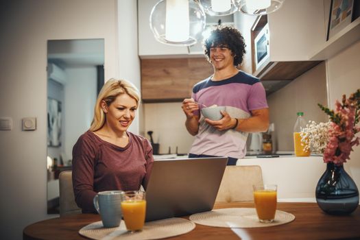Shot of a happy couple spending their morning time together and talking in the kitchen. They are using laptop and eating breakfast before work.