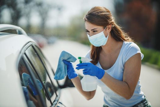 Cropped shot of a young smiling woman wearing protective mask busy wipes a her car outside with disinfectant spray.