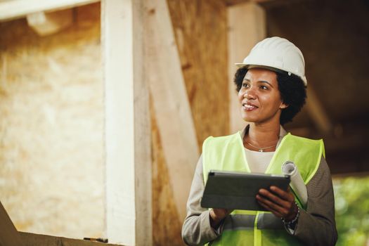 Shot of an African female architect using a digital tablet at the construction site of a new wooden house. She is wearing protective workwear and white helmet.
