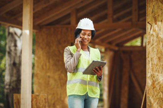 Shot of an African female architect using a smartphone and checking construction site of a new wooden house. She is wearing protective workwear and white helmet.