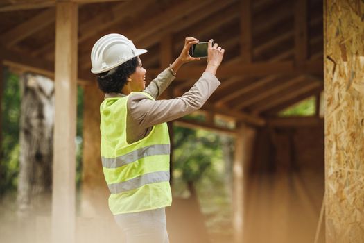 Shot of an African female architect taking photos on construction site  with smartphone during checking a new wooden house. She is wearing protective workwear and white helmet.