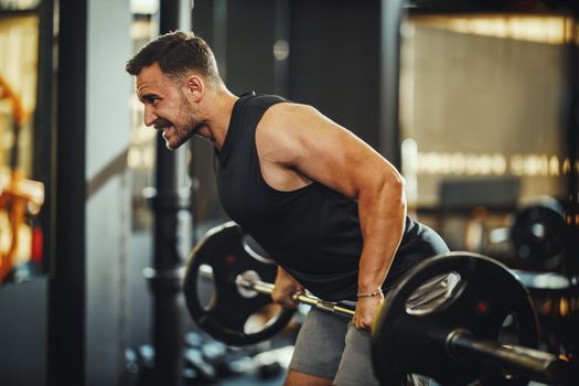Shot of a muscular guy in sportswear working out with barbell at the cross training gym. He is pumping up back muscule with heavy weight.