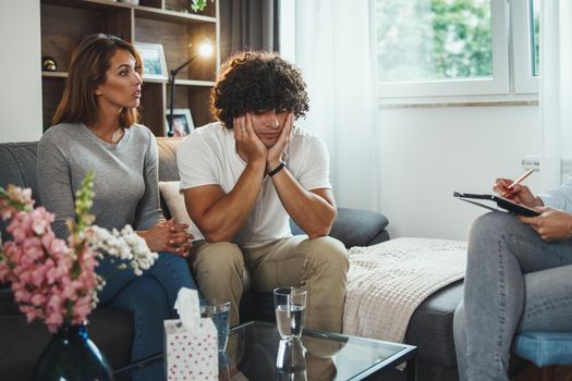Shot of a young couple during a counseling session with theirs female psychotherapist while being seated on a sofa inside of a living room.