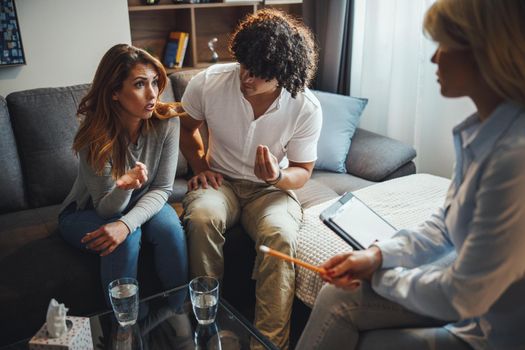 Shot of a young couple having a discussion during a counseling session with theirs female psychotherapist while being seated on a sofa inside of a living room.