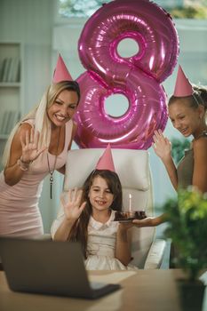 Beautiful young mom with her daugters at home during pandemic isolation have birthday party, she blowing candles and have conference call with friends and family.