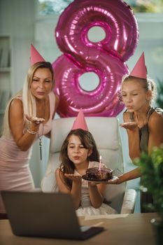 Beautiful young mom with her daugters at home during pandemic isolation have birthday party, she blowing candles and have conference call with friends and family.