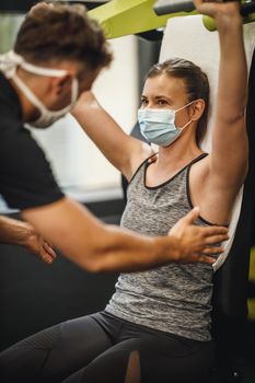 Shot of a muscular young woman with protective mask working out with personal trainer at the gym machine during Covid-19 pandemic. She is pumping up her shoulder muscule with heavy weight.