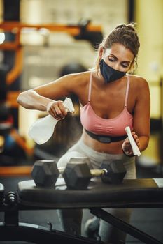 Shot of a muscular young woman with protective mask cleaning fitness gym equipment before workout during Covid-19 pandemic.
