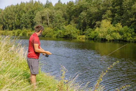 A man with a beard is fishing on the river. A fisherman with a fishing rod is fishing on the river bank.