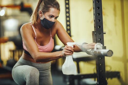 Shot of a muscular young woman with protective mask cleaning fitness gym equipment afther workout during Covid-19 pandemic.