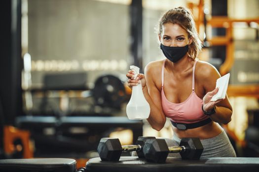 Shot of a muscular young woman with protective mask cleaning fitness gym equipment before workout during Covid-19 pandemic.