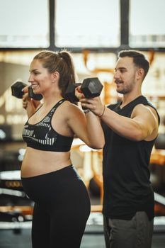 Shot of a pregnant woman working out with a dumbbell under control of the personal trainer at the gym.