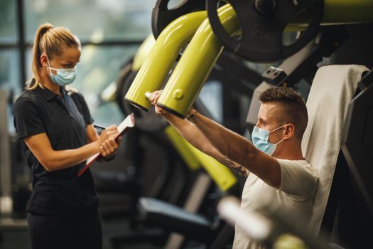 Shot of a muscular young woman with protective mask working out with personal trainer at the gym machine during Covid-19 pandemic. She is pumping up her chest muscule with heavy weight.