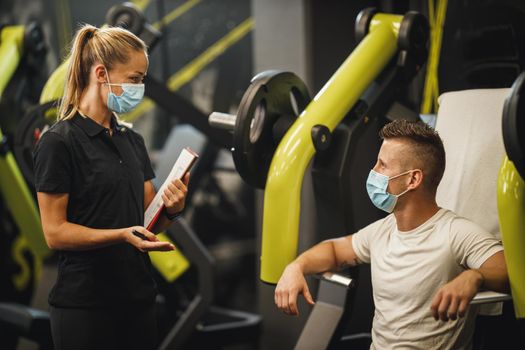 Shot of a muscular young woman with protective mask working out with personal trainer at the gym machine during Covid-19 pandemic. She is pumping up her chest muscule with heavy weight.