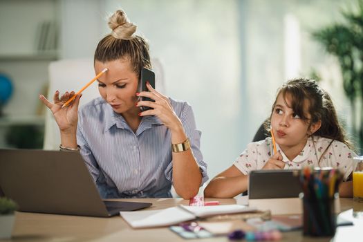 Overworked beautiful single mother using laptop while her daughter is learning at home during COVID-19 pandemic.