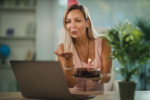 Alone attractive woman have birthday celebration at home during pandemic isolation and have video call with friends. She holding birthday cake with lighted candles.