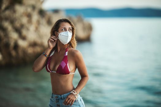 Shot of an attractive happy young woman with surgical mask enjoying a vacation on the beach during the COVID-19.