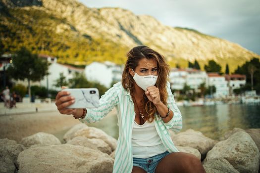 Shot of a young woman coughing during wearing a protective N95 mask and making video call with her smartphone while spending time on the seaside during the COVID-19.