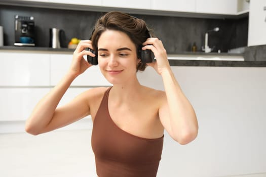 Smiling young fitness girl with headphones, exercise in her kitchen, indoor gym workout, listens music while doing pilates, training session from house.