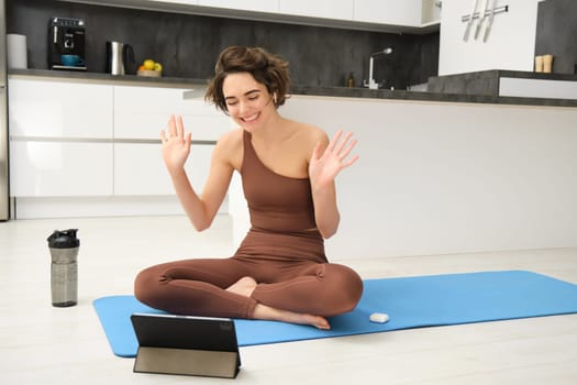 Smiling young fitness woman, instructor waves hands at tablet, records video blog about sport at home, says hello to workout class, stretching and doing gym exercises indoors.