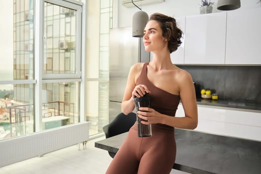 Portrait of smiling fitness girl, wearing sportswear for jogging, workout training, standing in kitchen and drinking water from sports bottle, looking fit and happy.