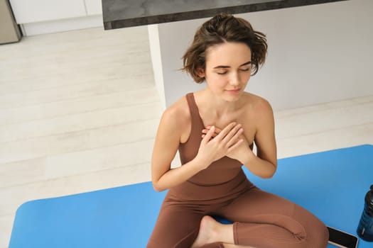 Lifestyle and sport concept. Upper angle shot of woman doing yoga, sitting on fitness mat at home, holds hands on chest, practice meditation with closed eyes and peaceful, relaxed smile.