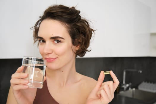 Dietry supplements and healthy lifestyle. Young woman taking vitamin C, D omega-3 with glass of water, standing in activewear, drinking after workout training in her kitchen.