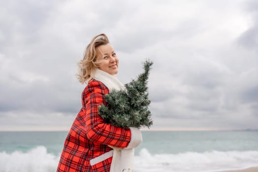 Blond woman holding Christmas tree by the sea. Christmas portrait of a happy woman walking along the beach and holding a Christmas tree in her hands. Dressed in a red coat, white dress