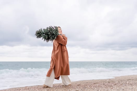 Blond woman Christmas sea. Christmas portrait of a happy woman walking along the beach and holding a Christmas tree on her shoulder. She is wearing a brown coat and a white suit