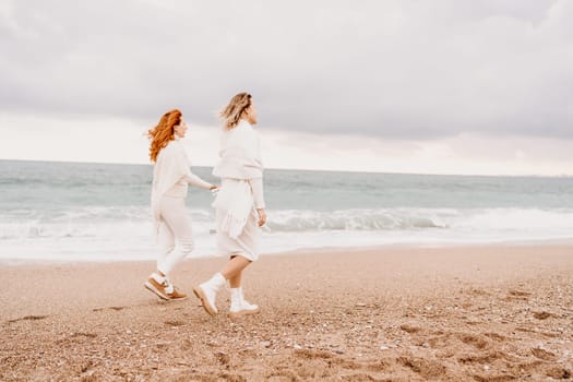 Women sea walk friendship spring. Two girlfriends, redhead and blonde, middle-aged walk along the sandy beach of the sea, dressed in white clothes. Against the backdrop of a cloudy sky and the winter sea. Weekend concept