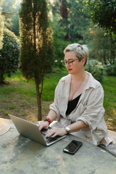 woman freelancer in glasses works at a computer at a white table in nature and spends her day productively.