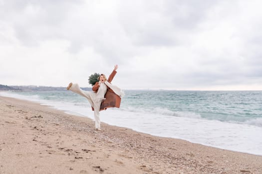 Blond woman Christmas sea. Christmas portrait of a happy woman walking along the beach and holding a Christmas tree on her shoulder. She is wearing a brown coat and a white suit