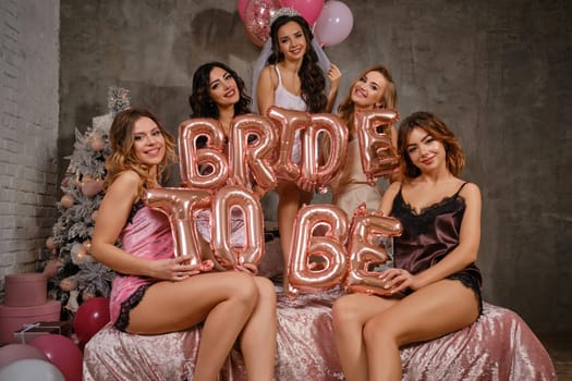 Pretty females in sexy lingerie and bride in veil, enjoying hen-party, sitting on bed, smiling, holding balloons in form of letters, built sentence bride to be. Christmas tree, decorations. Close-up.