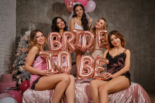 Pretty females in sexy lingerie and bride in veil, enjoying hen-party, sitting on bed, smiling, holding balloons in form of letters, built sentence bride to be. Christmas tree, decorations. Close-up.