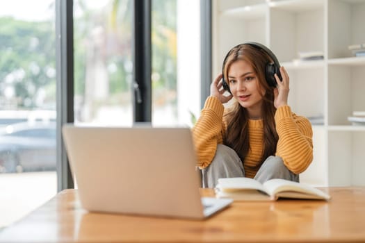 Asian college student at home using laptop attending online university classes listening with headphones.