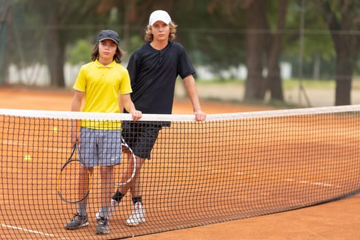 Two boys standing on the tennis court leaning on the net holding racquets and looking in the camera. Mid shot