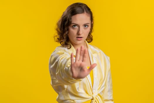 Young woman say No hold palm folded crossed hands in stop gesture, warning of finish, prohibited access, declining communication, body language, danger. Girl on yellow background. People lifestyles